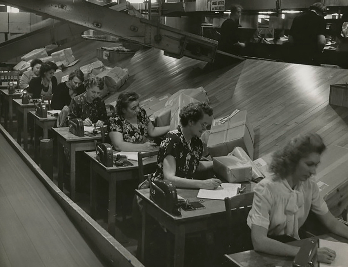 Black-and-white photo of a row of workers sitting at desks with packages.