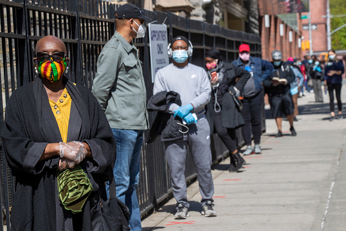 A line of people, all wearing masks, wait in line on a sidewalk.