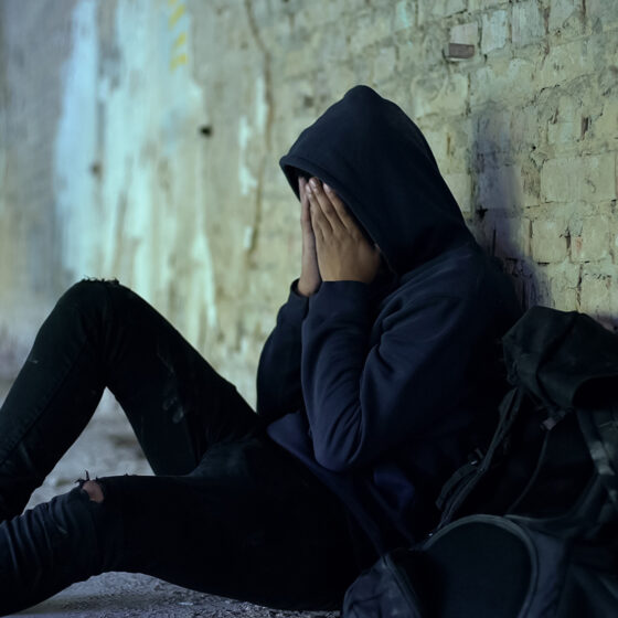 A teenager sits on the ground, leaned up against a brick wall with their hands on their face.