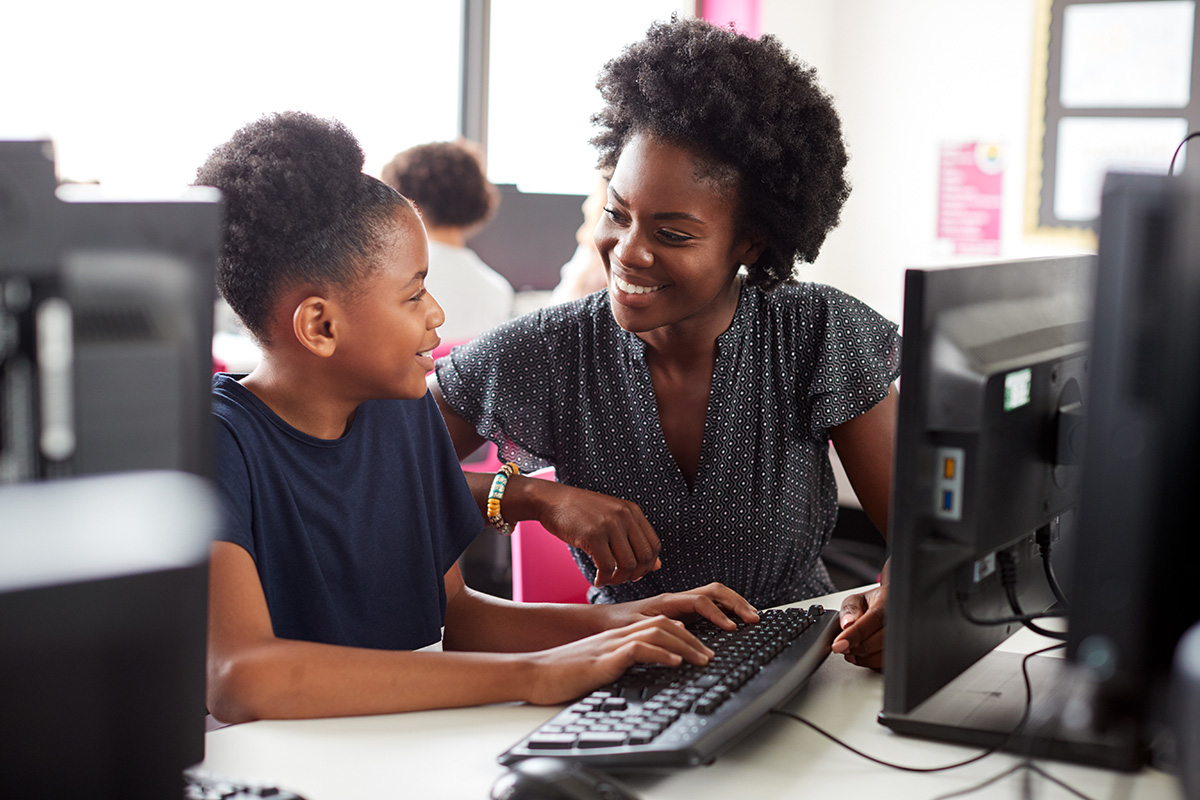 A Black youth is seen at a computer smiling at a Black teacher assisting her at a keyboard.
