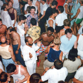Men in a variety of 80s style clothing some with their shirts off, are seen crowded on a busy dance floor.