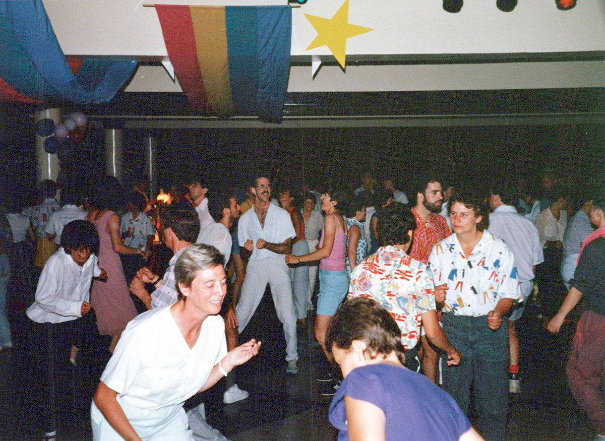 About 35 women and men dance in small groups on a checkered dance floor with coloured lights and banners hanging above their heads including a rainbow flag.