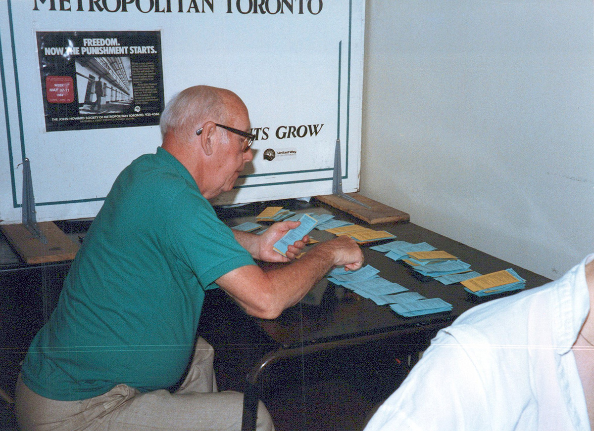 An older, white, presumably cisgender man in a green golf shirt is seen sitting at a desk sorting paper tickets into stacks.