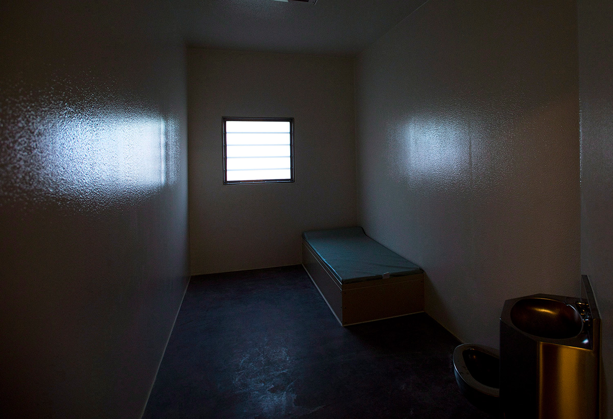 A small darkened cell with a small window, a narrow cot and a toilet.