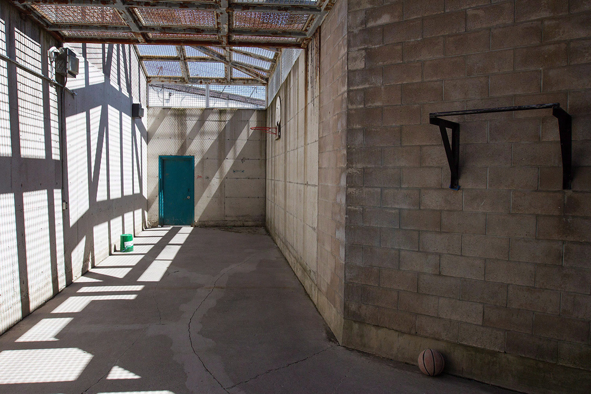 A small exercise yard with cinder wall and the sun shining from above through caging.