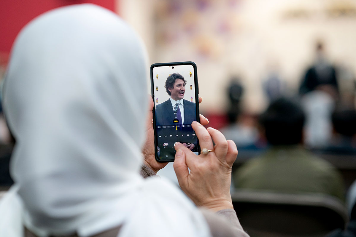 A woman in a white hijab adjusts the settings of her smartphone while capturing an image of a man with dark hair.