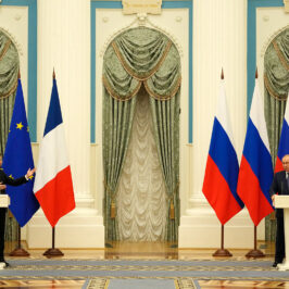 Two men stand far apart at podiums with Russian, EU and French flags behind them in an ornate hall.