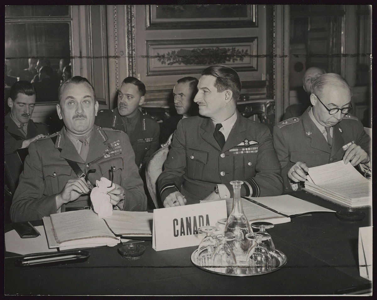 A black and white photo of men in military uniforms at a table with documents in front of them. A Canada sign also sits in front of them.