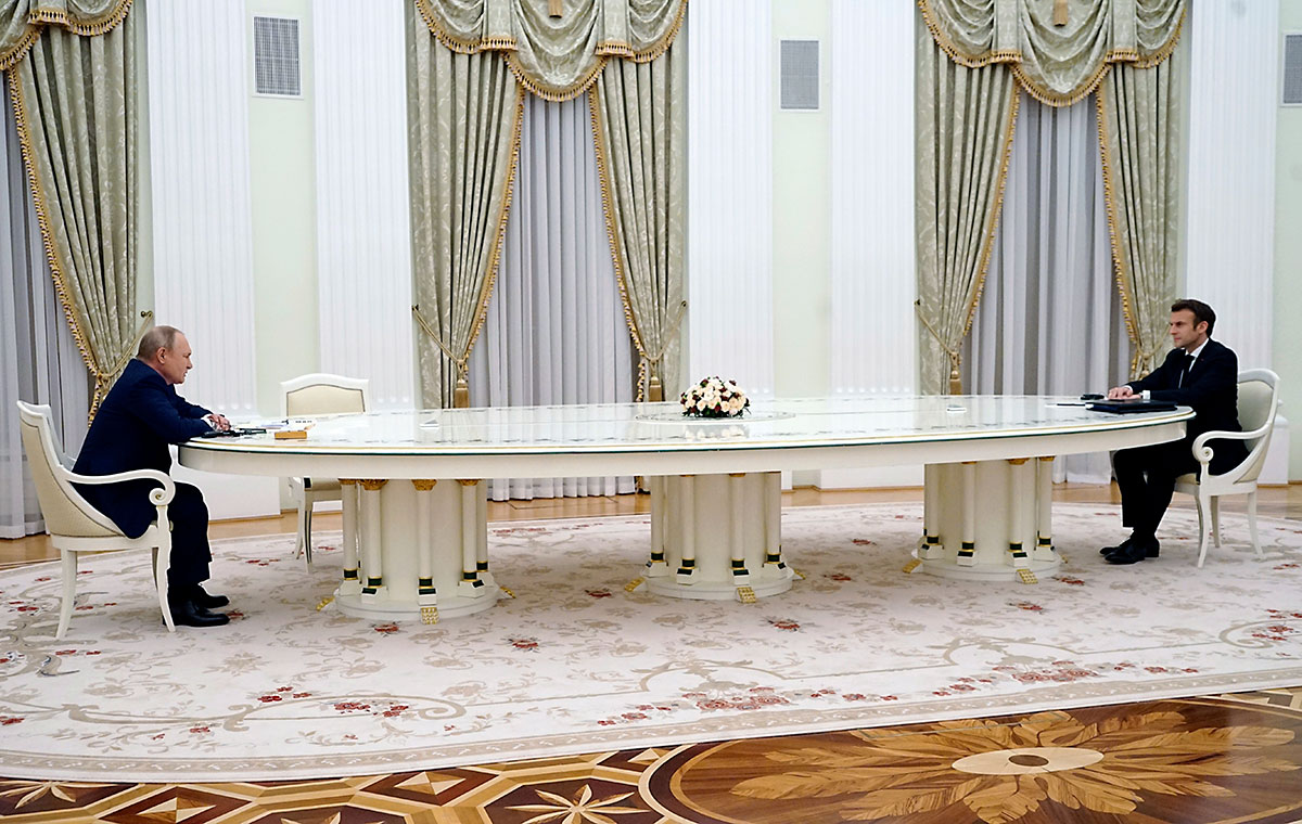 Two men sit far apart at opposite ends of a large, ornate white table in an ornate room with cream carpets and drapery.