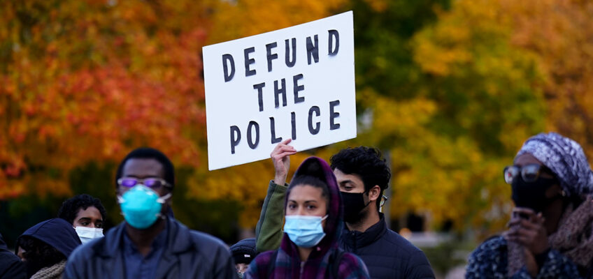 A protesters holds up a Defund the Police sign against a backdrop of autumn leaves.