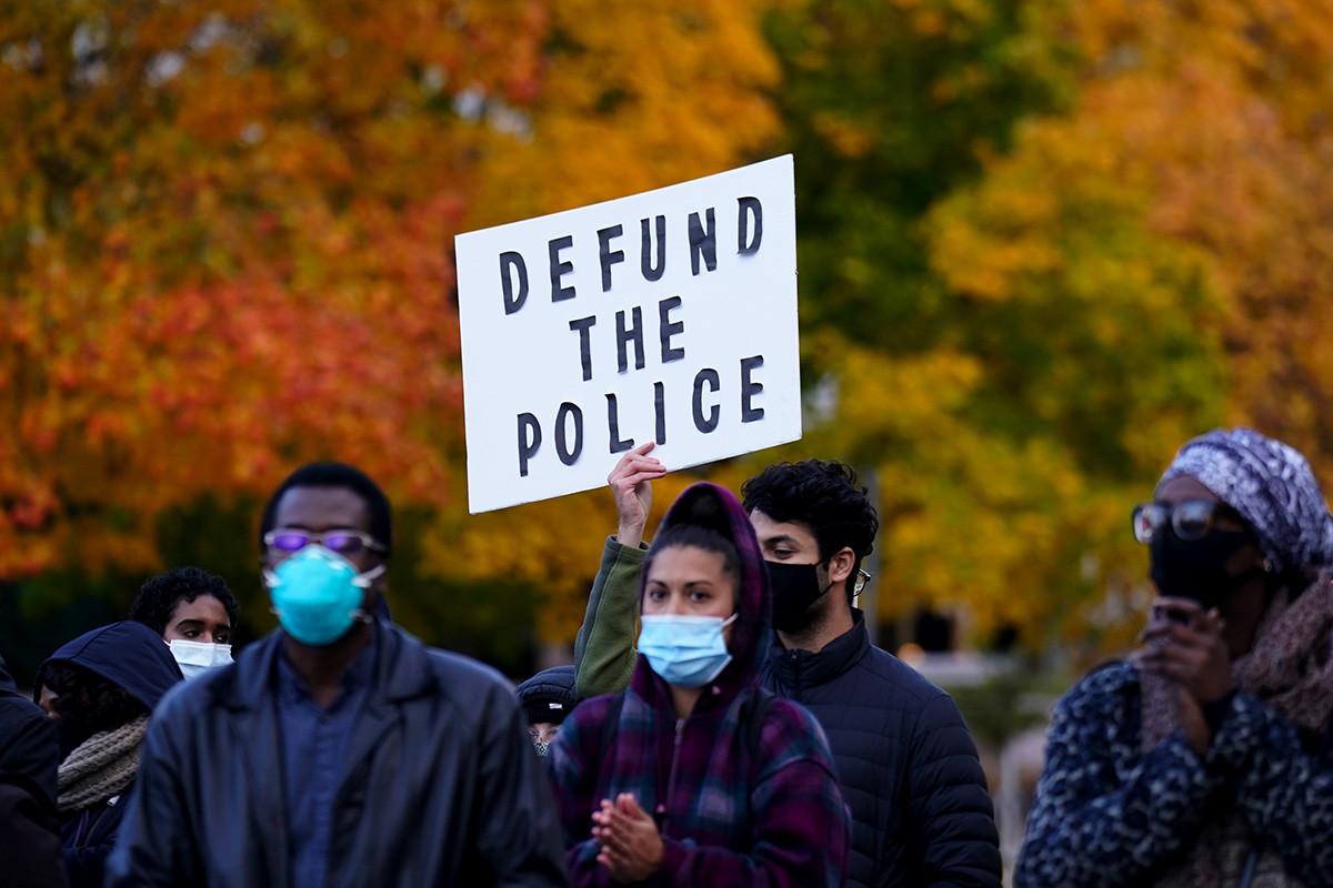 A protesters holds up a Defund the Police sign against a backdrop of autumn leaves.