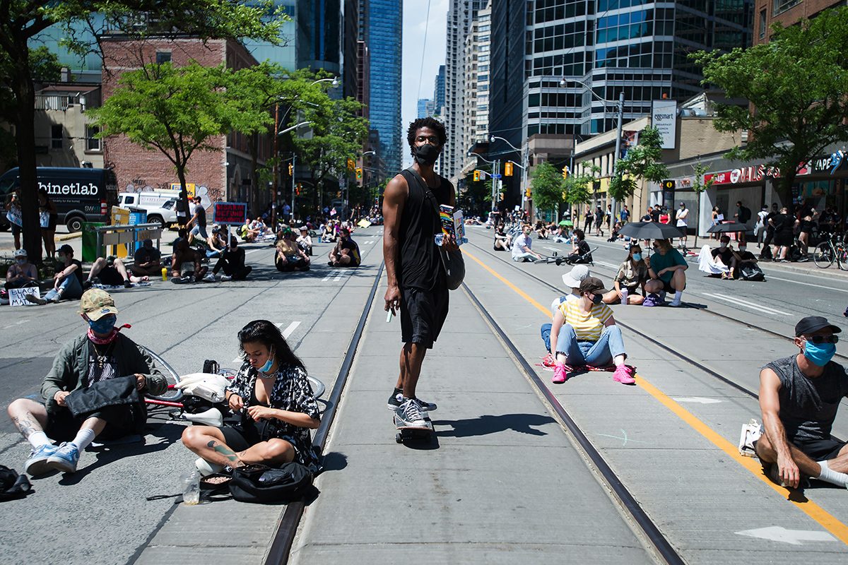 A Black man stands on a skateboard among people sitting on a city street during a protest.