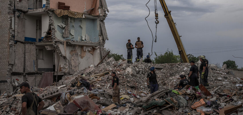 Rescue workers stand on the rubble of a destroyed apartment building. A couch is seen sitting a few storeys above them.