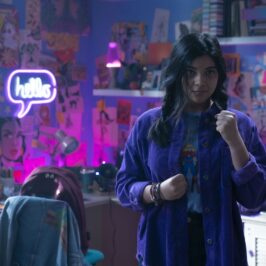 A young woman in her room surrounded by purple-hued walls and art and comic pictures, seen clenching one fist near her body and holding one up, looking forward.