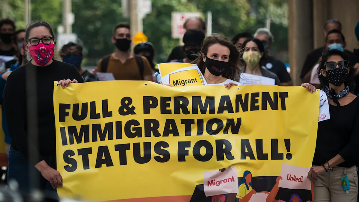 A woman wearing a face mask carries a large yellow sign with black lettering that reads: Full and permanent immigration status for all.