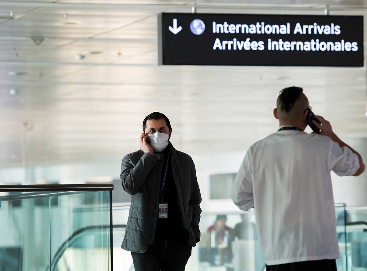 A man wearing a face mask chats on his mobile phone while standing below a sign that says 'International Arrivals' in an airport.