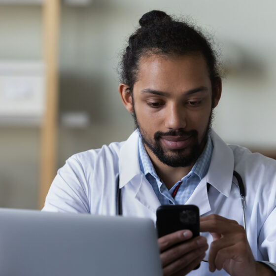 A man wearing a doctor coat sits behind a laptop, scrolling through his cellphone