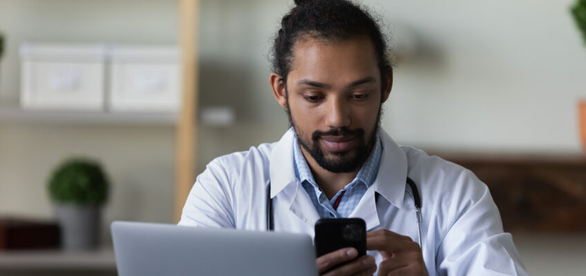 A man wearing a doctor coat sits behind a laptop, scrolling through his cellphone