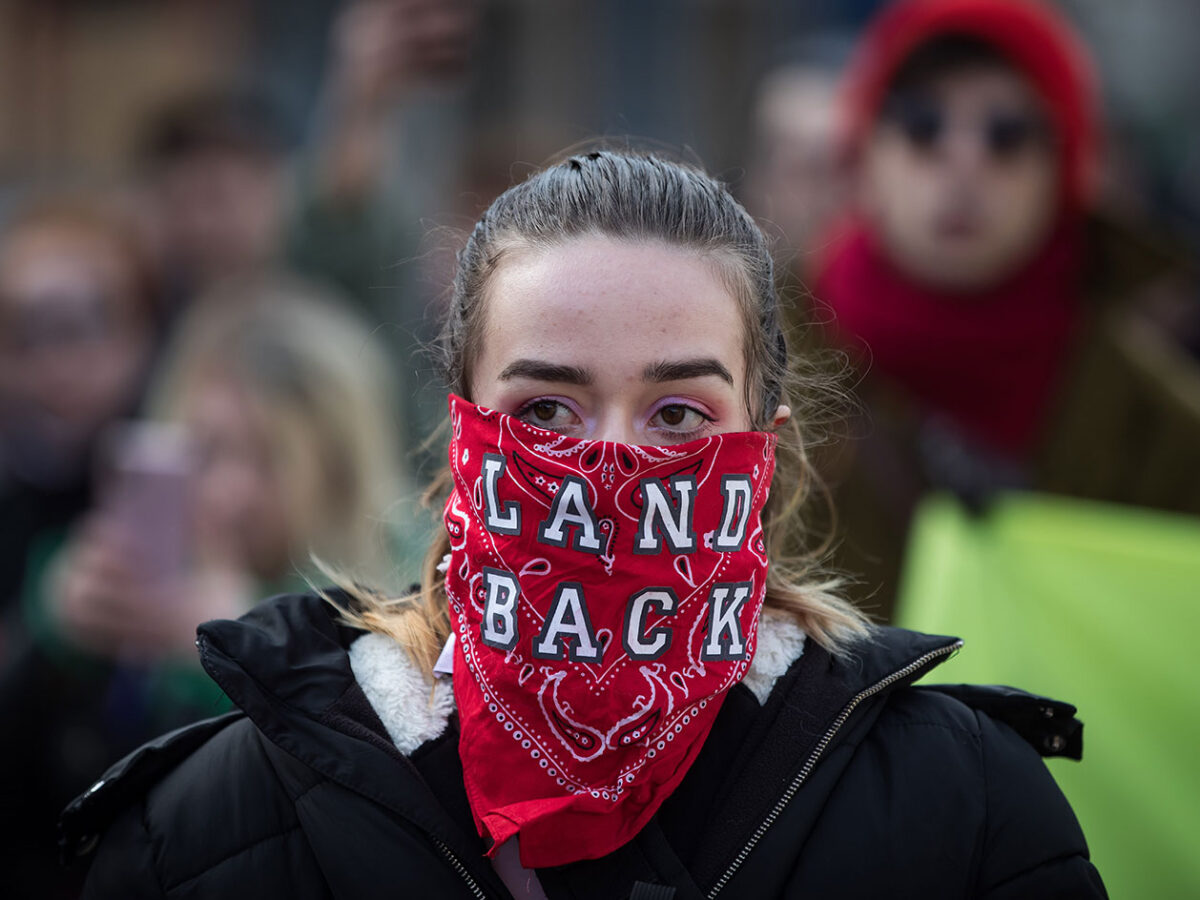 A woman wearing a red bandana over her face that reads: land back.