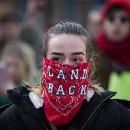 A woman wearing a red bandana over her face that reads: land back.
