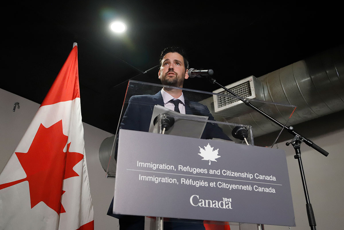 A man speaks from behind a 'Immigration, Refugees and Citizenship Canada' branded podium. A Canadian flag stands in the background.