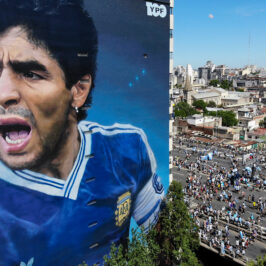A large mural of Diego Maradona on the side of a building. People carrying Argentine flags walk past.