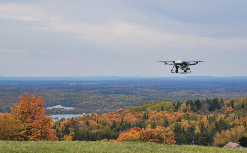 A remotely piloted long-range survey drone flies above an expansive area of deciduous and coniferous trees in autumn and some small bodies of water with a blue sky