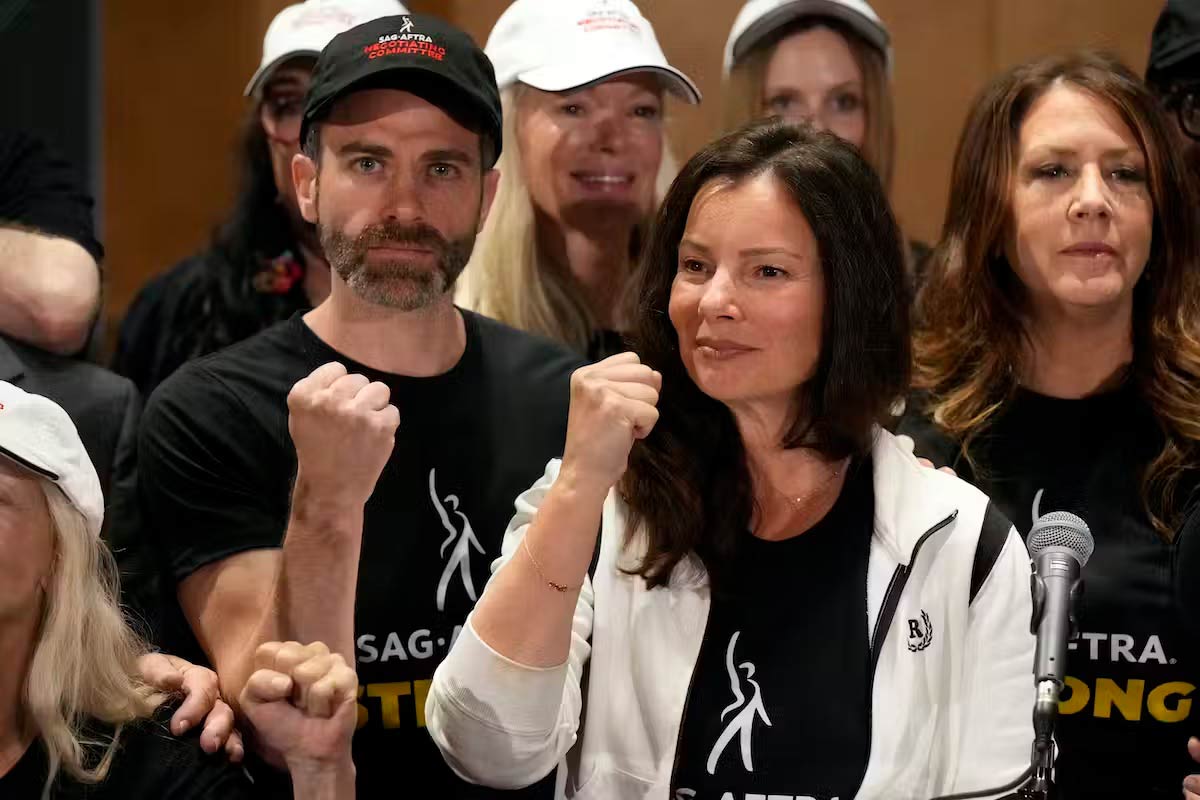 A woman and a crowd of people wearing SAG-AFTRA shirts hold their fists up