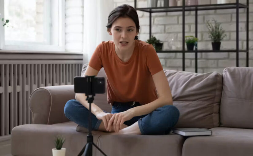 A young woman sits crosslegged on a couch videotaping herself for a social media post.