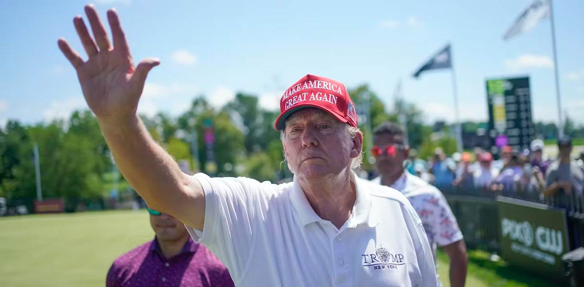 A man in a white golf shirt wearing a Make America Great Again red ballcap waves without smiling.