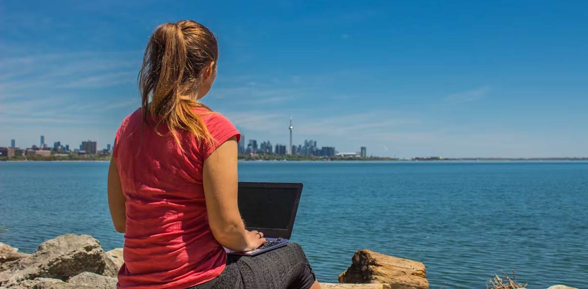 A woman working on a laptop sits by a lake. The Toronto skyline is in the distance.