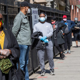 A line of people, all wearing masks, wait in line on a sidewalk.
