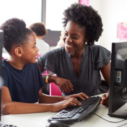 A Black youth is seen at a computer smiling at a Black teacher assisting her at a keyboard.