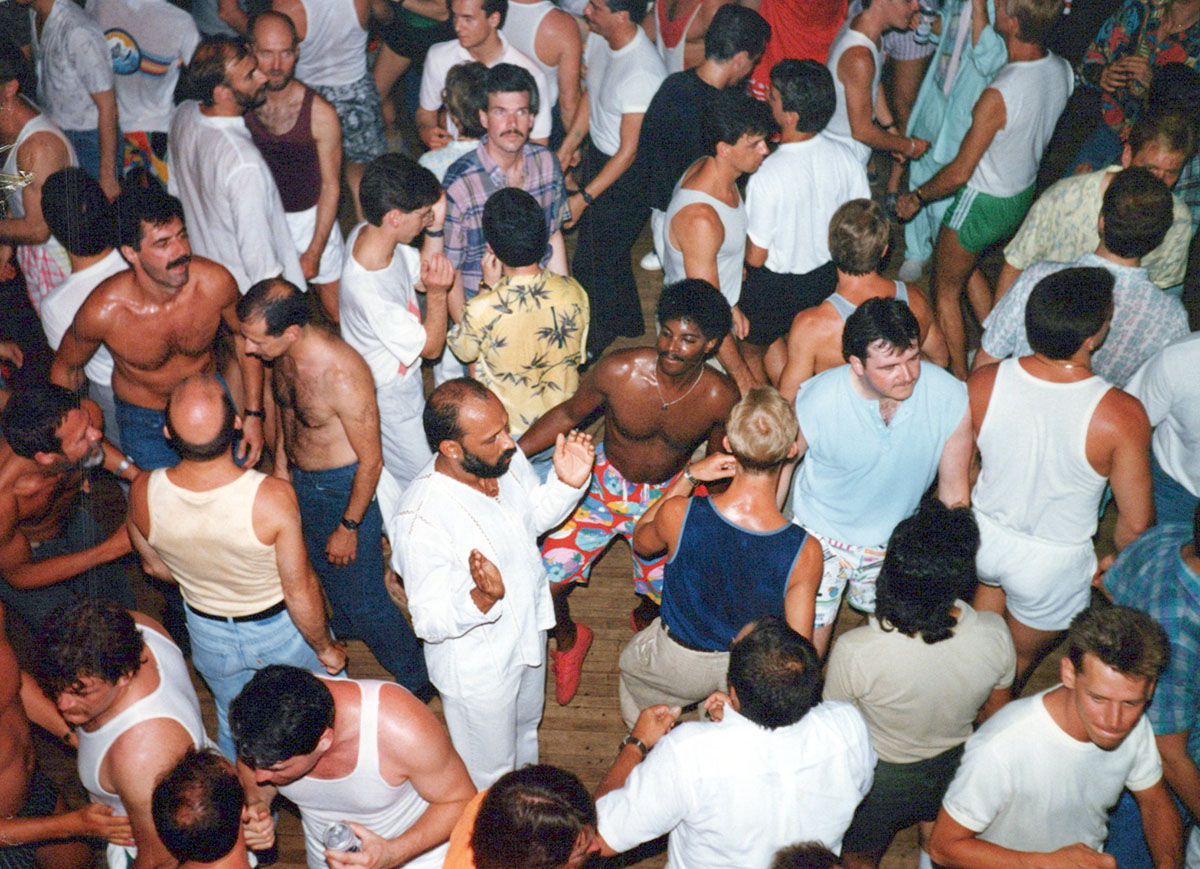 Men in a variety of 80s style clothing some with their shirts off, are seen crowded on a busy dance floor.