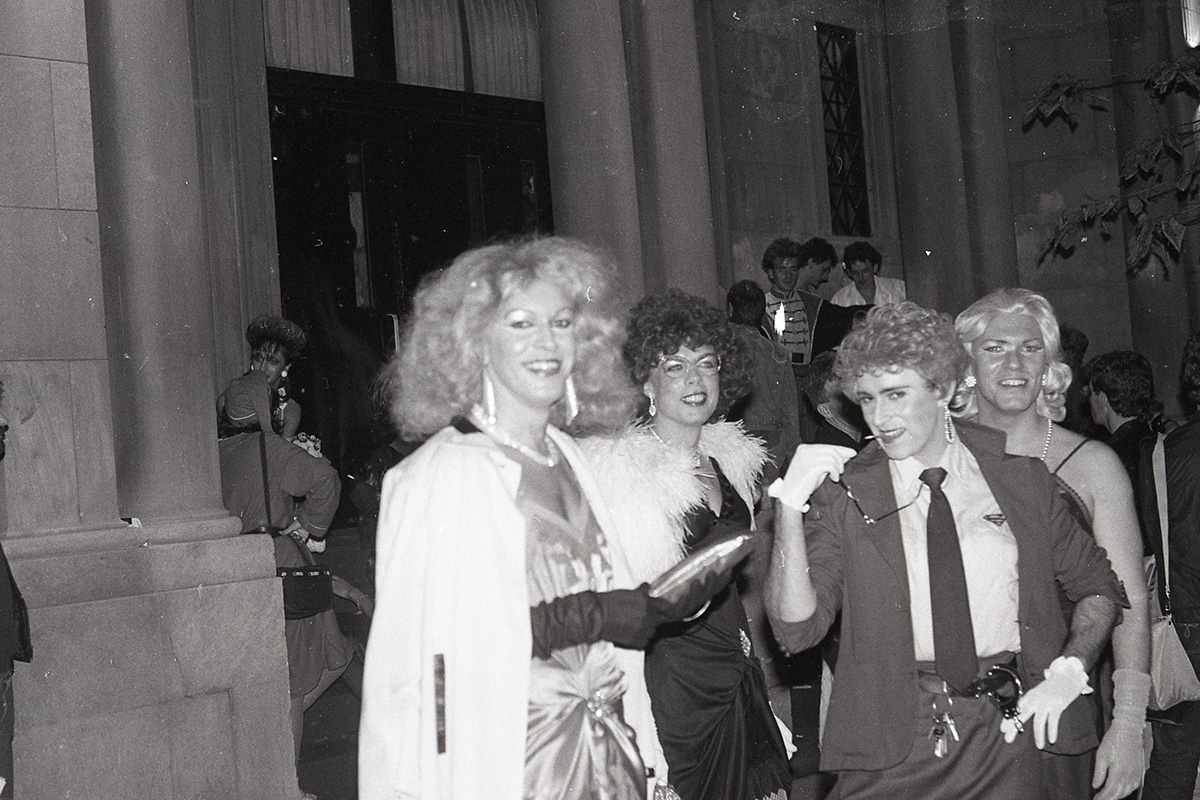 Four white men in drag smile while looking at the camera outside of the Masonic Temple where GCDC dances were held. There is a large group of people behind the four men.
