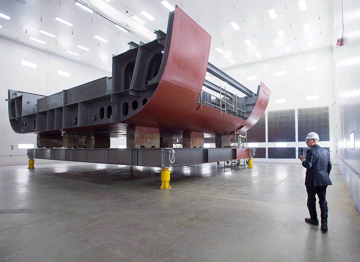 A man walks past a large component of a ship inside a shipbuilding facility.
