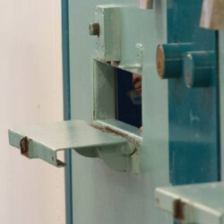 A sliver of the face of an inmate is seen inside a tiny slot in a prison cell door.