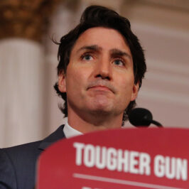 A dark haired man stands behind microphone and a podium that reads 'tougher gun laws'.