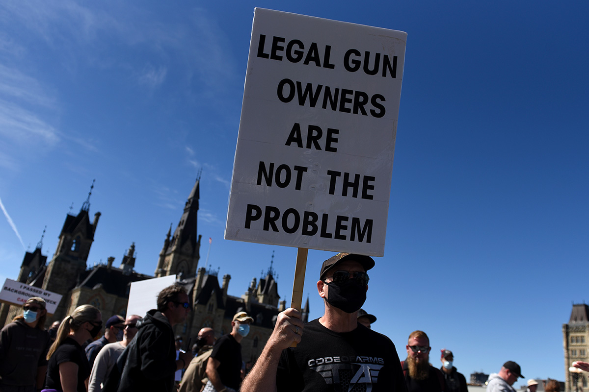 A man wearing a mask holds a sign that reading Legal Gun Owners Are Not The Problem.