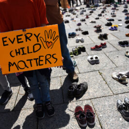 Dozens of pairs of children's shoes on the pavement. A person in an orange T-shirt stands beside the shoes with a sign that reads Every Child Matters.