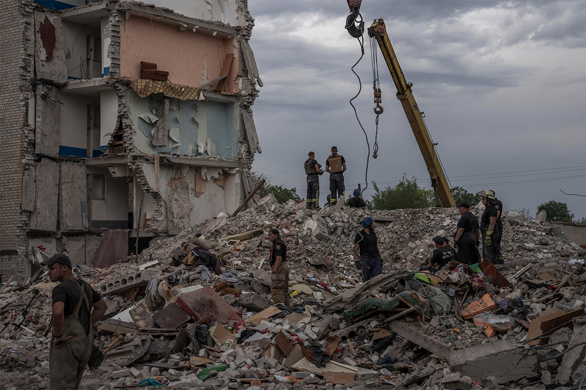 Rescue workers stand on the rubble of a destroyed apartment building. A couch is seen sitting a few storeys above them.