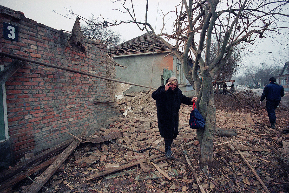 An elderly woman weeps as she stands near rubble with her hand on a tree.