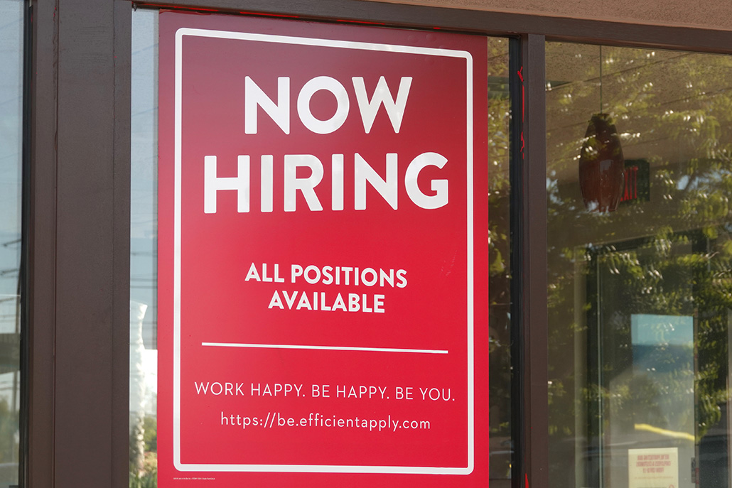 A 'Now Hiring' sign hanging in the window of a building.