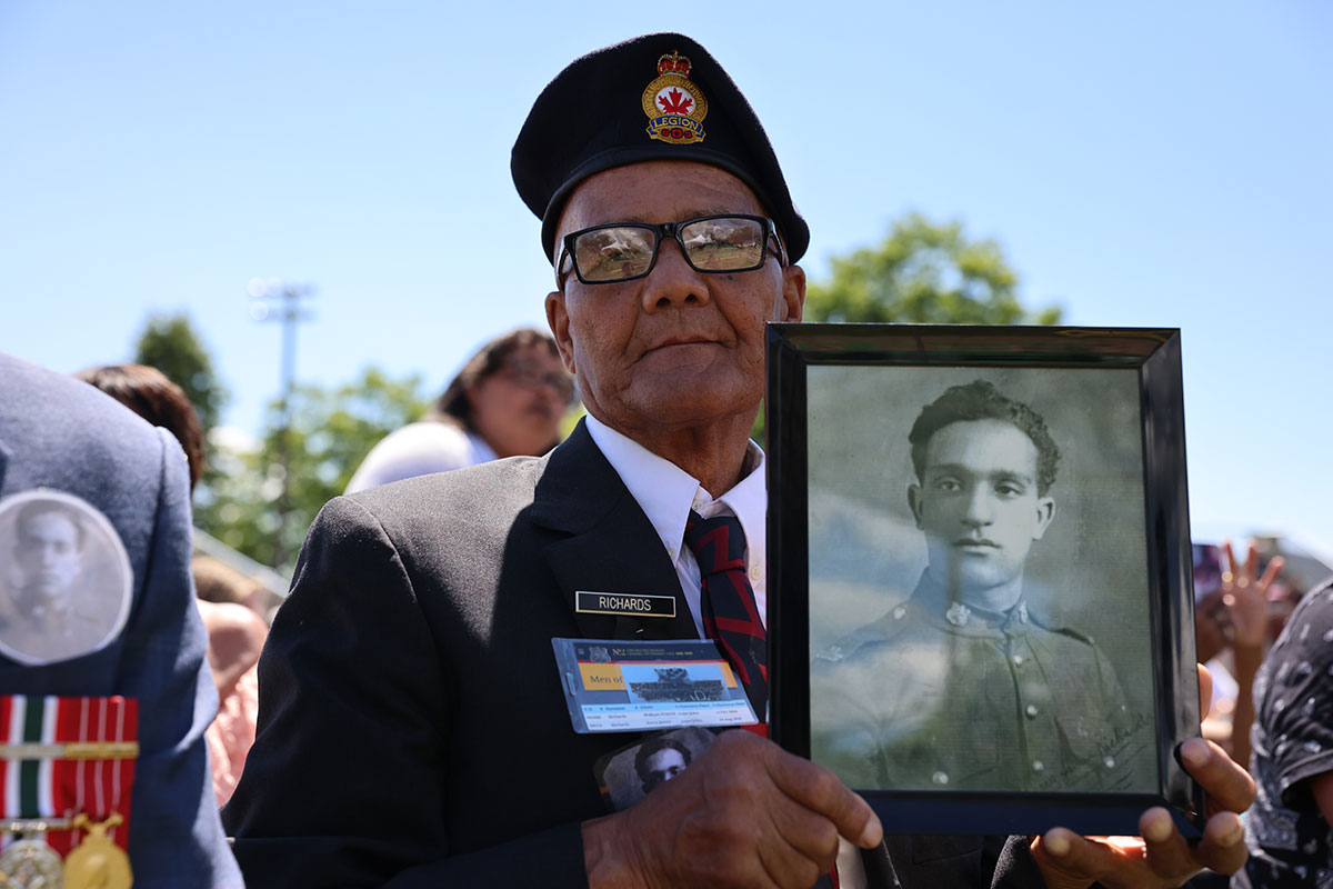 An elderly man in military uniform holds a black and white photo of a younger man also in uniform