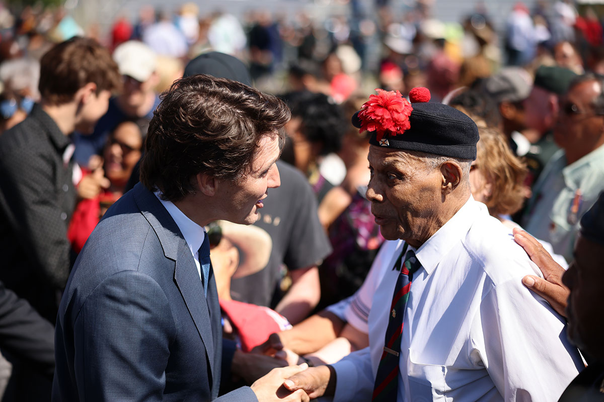 A white man in a dark suit shakes hands with an elderly black man wearing a shirt and tie and military cap.
