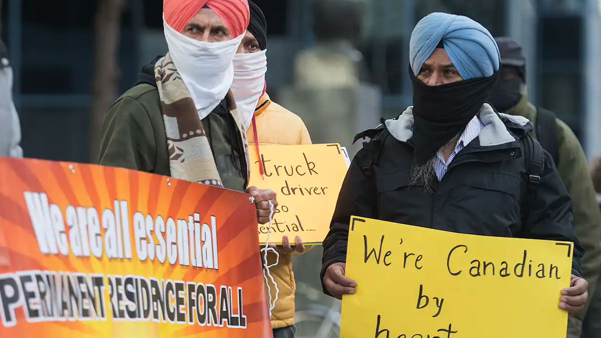 Men wearing face masks and Sikh turbans hold signs that read: we are all essential, permanent residence for all.