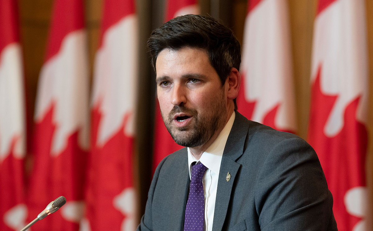 A man in a suit and tie speaking into a microphone. Behind him stands a line of Canadian flags.