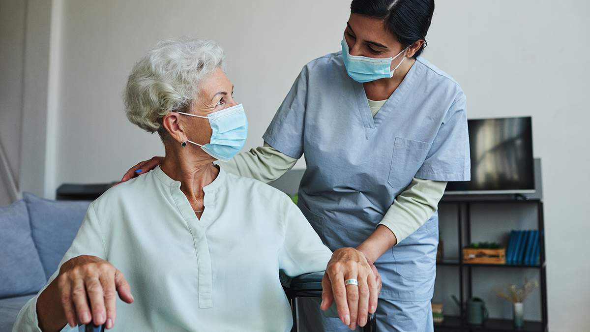 A female nurse assists a senior woman in wheelchair. Both are wearing masks.