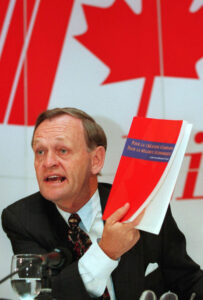 A man siting in front of a Canadian flag hold up a red book.