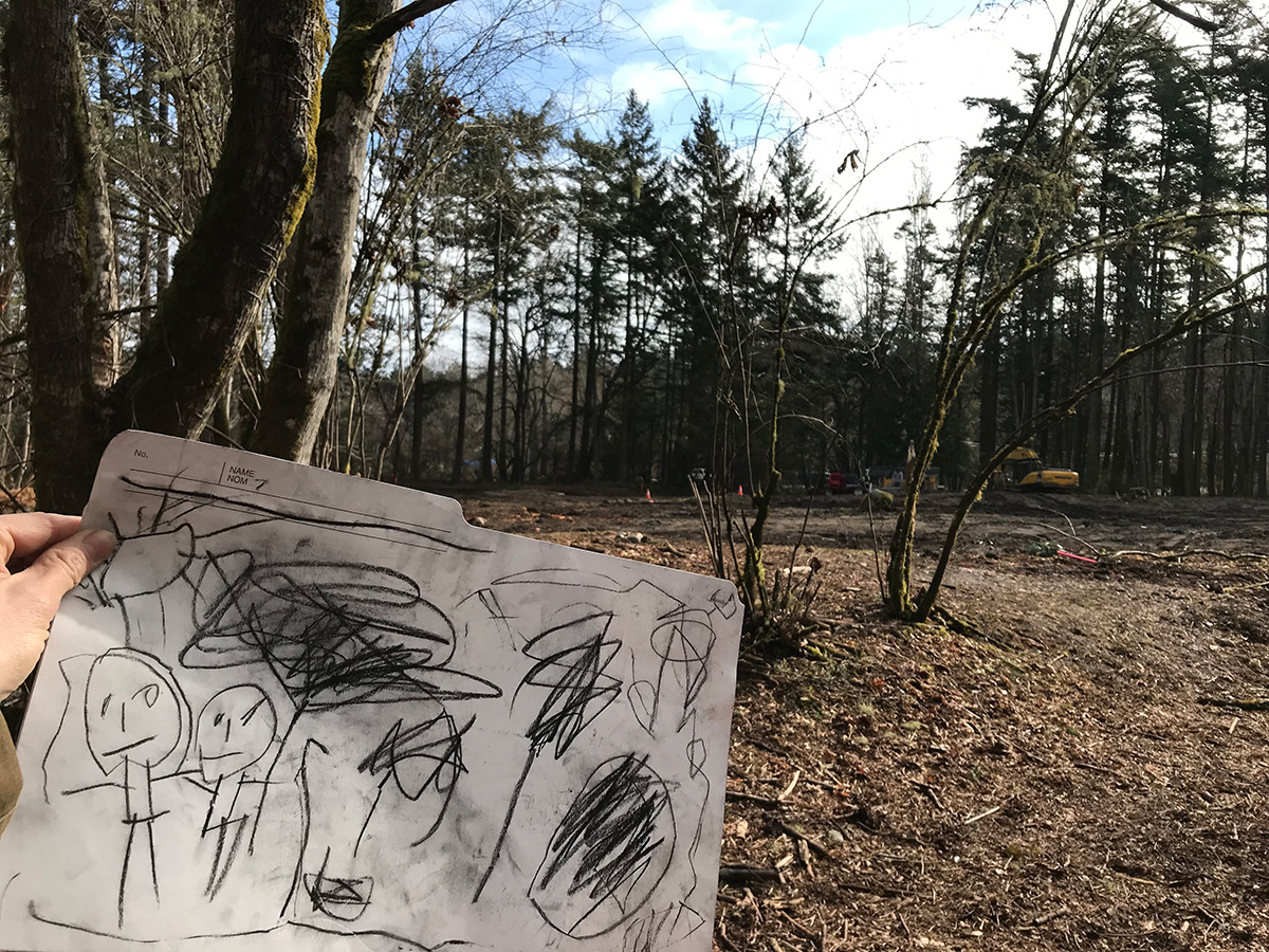 A hand is seen holding a child's drawing in front of woods.